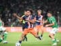 Players in green and navy kits chase the ball on a soccer pitch during a match, with a central player in orange shorts reaching for control.