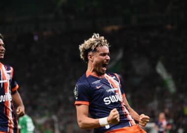 Two soccer players in blue and orange striped jerseys celebrate on the field, fists clenched and mouths open.