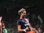 Two soccer players in blue and orange striped jerseys celebrate on the field, fists clenched and mouths open.