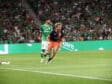Two soccer players chase the ball on a green pitch while a crowd in green fills the stands; one in green kit, one in dark blue and orange lunges forward.
