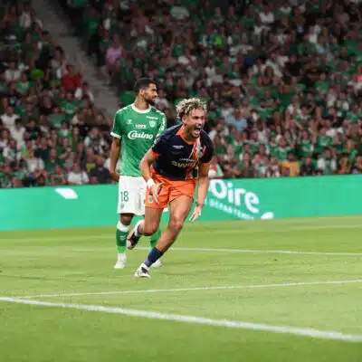 Two soccer players chase the ball on a green pitch while a crowd in green fills the stands; one in green kit, one in dark blue and orange lunges forward.