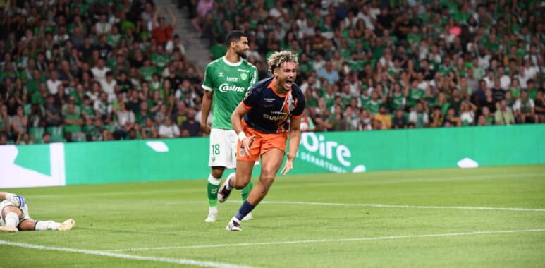 Two soccer players chase the ball on a green pitch while a crowd in green fills the stands; one in green kit, one in dark blue and orange lunges forward.