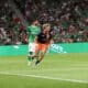 Two soccer players chase the ball on a green pitch while a crowd in green fills the stands; one in green kit, one in dark blue and orange lunges forward.