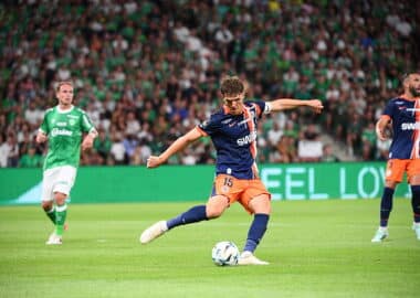 Soccer player in blue and orange kit kicks the ball during a match, with a green-clad opponent and a packed crowd in the background.