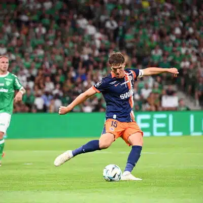 Soccer player in blue and orange kit kicks the ball during a match, with a green-clad opponent and a packed crowd in the background.