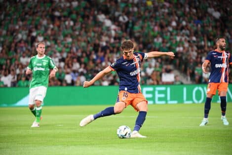 Soccer player in blue and orange kit kicks the ball during a match, with a green-clad opponent and a packed crowd in the background.