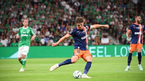 Soccer player in blue and orange kit kicks the ball during a match, with a green-clad opponent and a packed crowd in the background.
