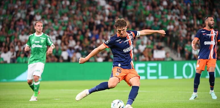Soccer player in blue and orange kit kicks the ball during a match, with a green-clad opponent and a packed crowd in the background.