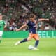 Soccer player in blue and orange kit kicks the ball during a match, with a green-clad opponent and a packed crowd in the background.