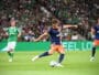 Soccer player in blue and orange kit kicks the ball during a match, with a green-clad opponent and a packed crowd in the background.