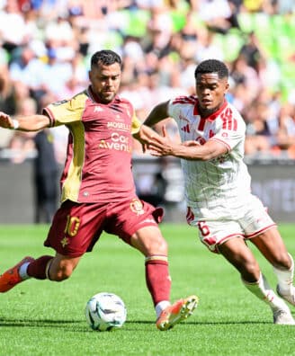 Two soccer players chase the ball during a daytime match, one in maroon and gold and the other in white with red stripes, goal in sighted crowd backdrop.