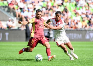 Two soccer players chase the ball during a daytime match, one in maroon and gold and the other in white with red stripes, goal in sighted crowd backdrop.