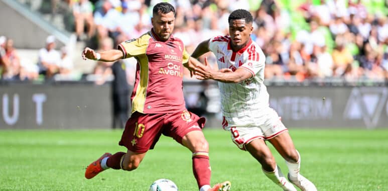 Two soccer players chase the ball during a daytime match, one in maroon and gold and the other in white with red stripes, goal in sighted crowd backdrop.