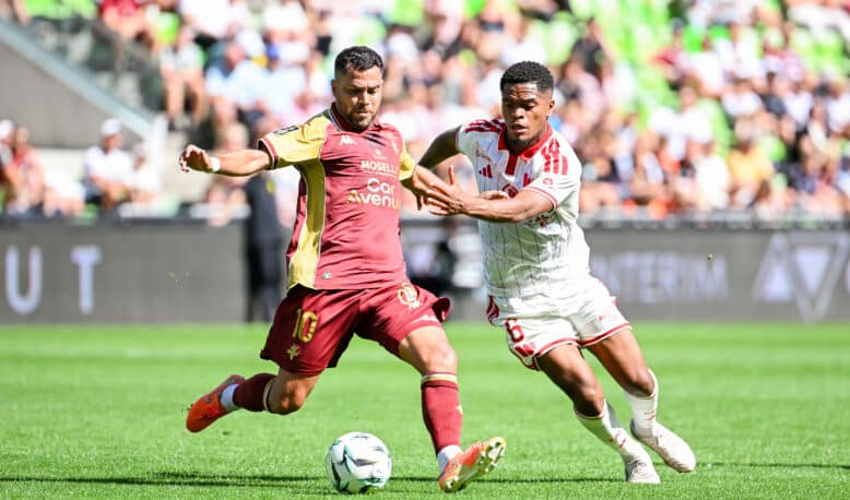 Two soccer players chase the ball during a daytime match, one in maroon and gold and the other in white with red stripes, goal in sighted crowd backdrop.