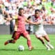Two soccer players chase the ball during a daytime match, one in maroon and gold and the other in white with red stripes, goal in sighted crowd backdrop.