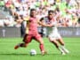 Two soccer players chase the ball during a daytime match, one in maroon and gold and the other in white with red stripes, goal in sighted crowd backdrop.