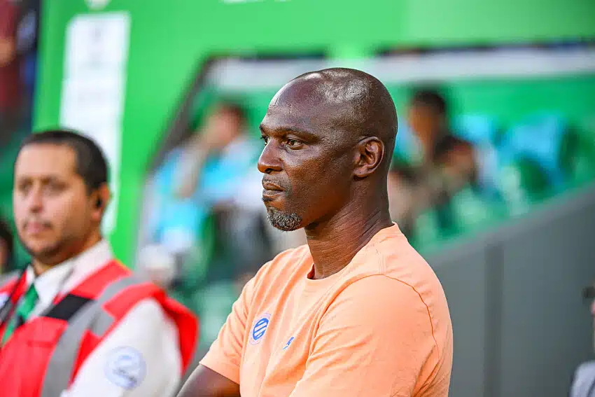 African man in a peach shirt sits with folded arms, watching a game in a stadium.