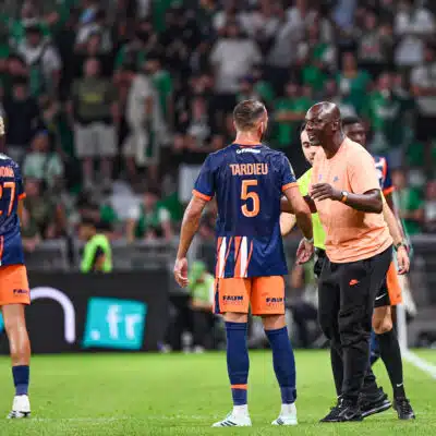 Soccer players in blue&ndash;orange uniforms on a green pitch sharing a moment as a referee speaks nearby, crowded stands in the background.