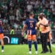 Soccer players in blue&ndash;orange uniforms on a green pitch sharing a moment as a referee speaks nearby, crowded stands in the background.