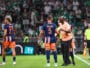 Soccer players in blue&ndash;orange uniforms on a green pitch sharing a moment as a referee speaks nearby, crowded stands in the background.