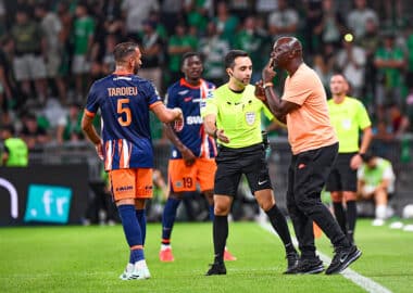 Soccer referee in a neon green shirt talks with a player in an orange-blue kit while teammates watch on a green pitch.