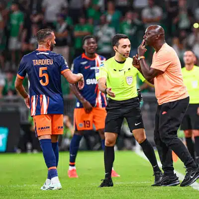 Soccer referee in a neon green shirt talks with a player in an orange-blue kit while teammates watch on a green pitch.
