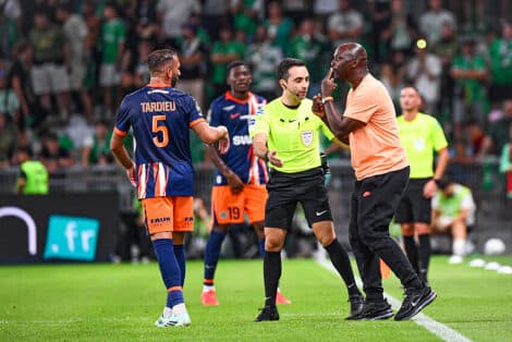 Soccer referee in a neon green shirt talks with a player in an orange-blue kit while teammates watch on a green pitch.