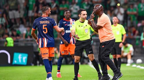 Soccer referee in a neon green shirt talks with a player in an orange-blue kit while teammates watch on a green pitch.