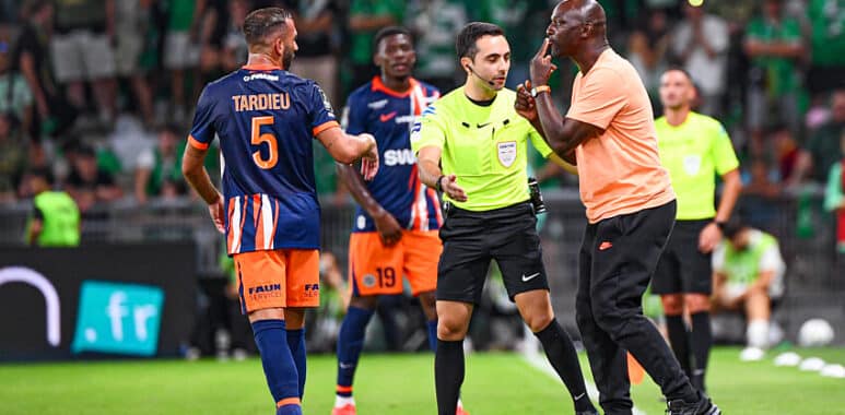 Soccer referee in a neon green shirt talks with a player in an orange-blue kit while teammates watch on a green pitch.