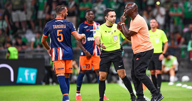 Soccer referee in a neon green shirt talks with a player in an orange-blue kit while teammates watch on a green pitch.