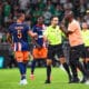 Soccer referee in a neon green shirt talks with a player in an orange-blue kit while teammates watch on a green pitch.