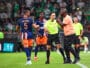 Soccer referee in a neon green shirt talks with a player in an orange-blue kit while teammates watch on a green pitch.