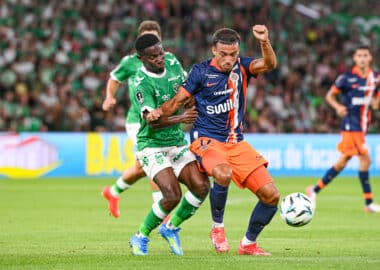Two soccer players from opposing teams chase the ball during a match on a green field, with a crowded stadium in the background.