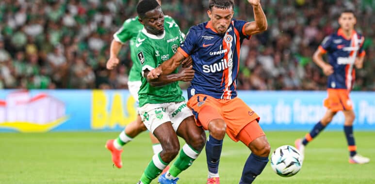 Two soccer players from opposing teams chase the ball during a match on a green field, with a crowded stadium in the background.