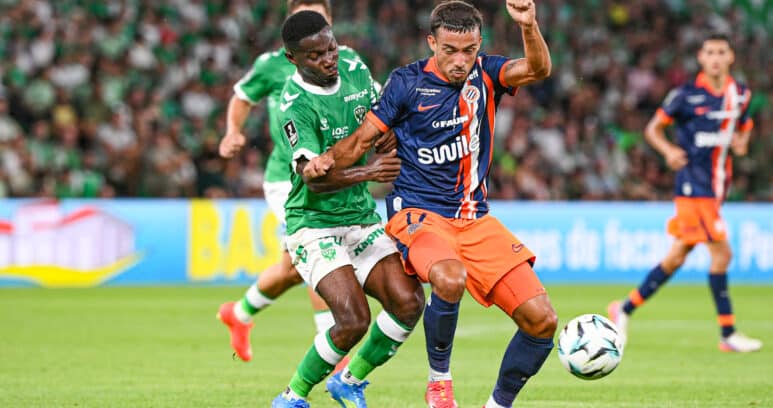 Two soccer players from opposing teams chase the ball during a match on a green field, with a crowded stadium in the background.