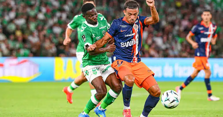 Two soccer players clash for the ball during a match, one in green and white and the other in navy and orange, on a grassy field with a blurred crowd in the background.