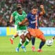 Two soccer players clash for the ball during a match, one in green and white and the other in navy and orange, on a grassy field with a blurred crowd in the background.