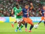 Two soccer players clash for the ball during a match, one in green and white and the other in navy and orange, on a grassy field with a blurred crowd in the background.