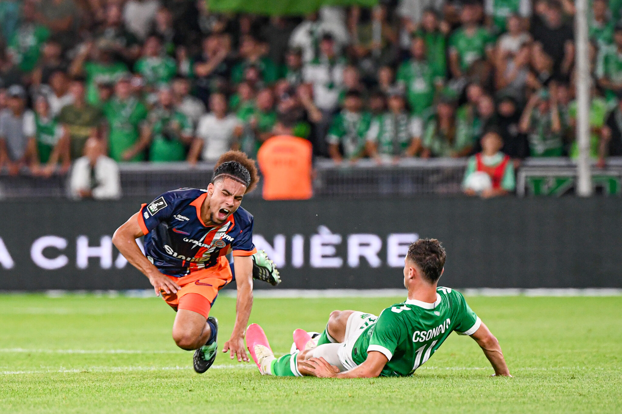 Abderrebi AS Saint-Etienne v Montpellier Herault SC - Ligue 2 BKT 20262027 | AllezPaillade.com Soccer match action: player in orange and navy shoots the ball while a green-clad goalkeeper slides in to block near the goal, crowd blurred in background.
