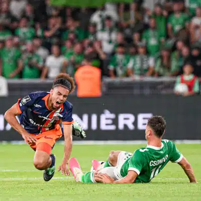 Soccer match action: player in orange and navy shoots the ball while a green-clad goalkeeper slides in to block near the goal, crowd blurred in background.