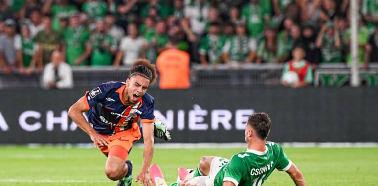 Soccer match action: player in orange and navy shoots the ball while a green-clad goalkeeper slides in to block near the goal, crowd blurred in background.