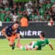 Soccer match action: player in orange and navy shoots the ball while a green-clad goalkeeper slides in to block near the goal, crowd blurred in background.