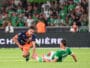 Soccer match action: player in orange and navy shoots the ball while a green-clad goalkeeper slides in to block near the goal, crowd blurred in background.