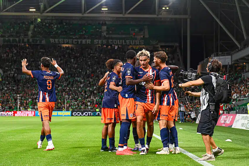 Soccer players in blue and orange jerseys hug and celebrate on the pitch as a photographer films nearby.