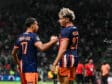 Two soccer players in navy and orange uniforms shake hands on the field as a match continues, with a camera to the right and a blurred crowd in the stands behind them.