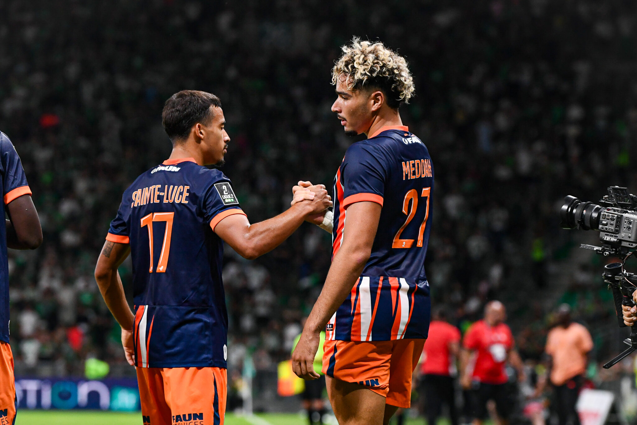 Sainte-Luce Meddah AS Saint-Etienne v Montpellier Herault SC - Ligue 2 BKT 20262027 | AllezPaillade.com Two soccer players in navy and orange uniforms shake hands on the field as a match continues, with a camera to the right and a blurred crowd in the stands behind them.