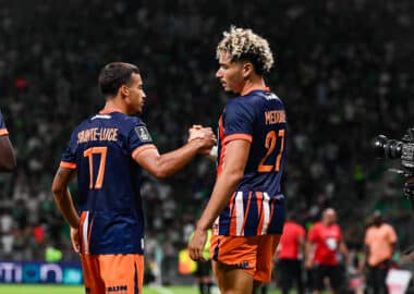 Two soccer players in navy and orange uniforms shake hands on the field as a match continues, with a camera to the right and a blurred crowd in the stands behind them.