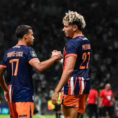 Two soccer players in navy and orange uniforms shake hands on the field as a match continues, with a camera to the right and a blurred crowd in the stands behind them.