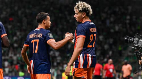Two soccer players in navy and orange uniforms shake hands on the field as a match continues, with a camera to the right and a blurred crowd in the stands behind them.
