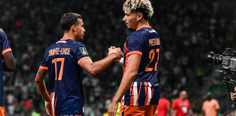 Two soccer players in navy and orange uniforms shake hands on the field as a match continues, with a camera to the right and a blurred crowd in the stands behind them.